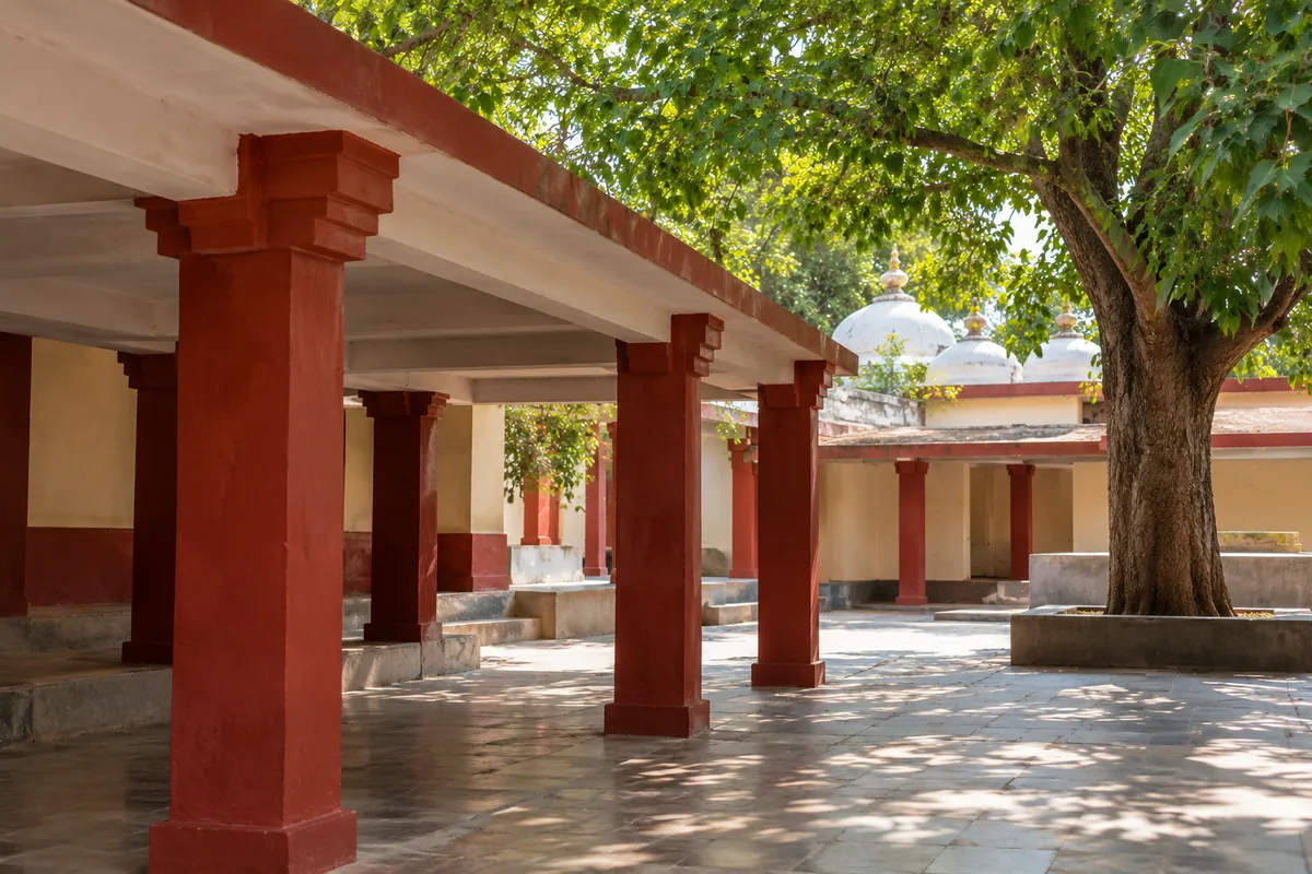 White and red painted gateway and low domed temple buildings beneath shade trees at Maa Majhighariani Temple