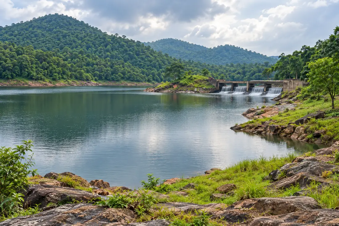 A quiet reservoir and low spillway sit among forested hills at Pitamahal